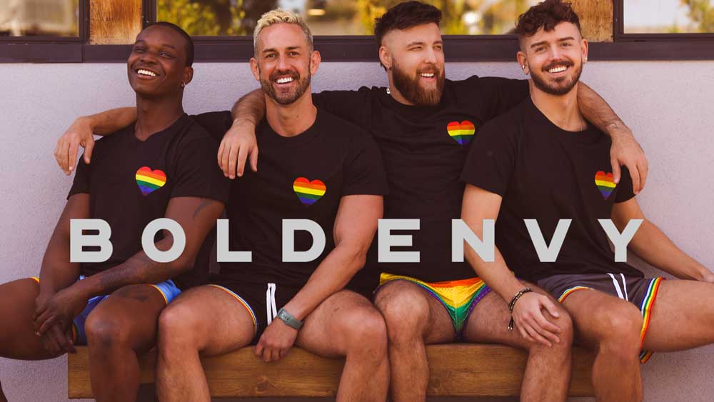 Four men sitting on a bench wearing black t-shirts with rainbow flags and colorful shorts, with 'BOLD ENVY' text overlay.
